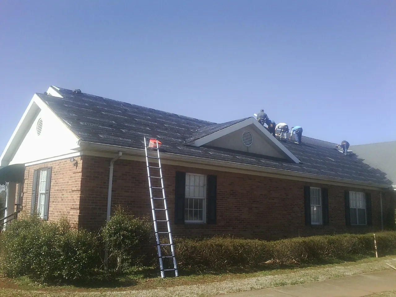 Crew tearing shingles off a brick ranch roof with a ladder set — Emergency Roof Repair work by our crew in Noonan