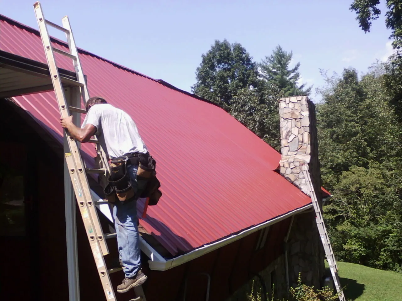 Roofer on a ladder beside a red metal roof and stone chimney — Soffit & Fascia Repair by Roofing Brothers in Noonan